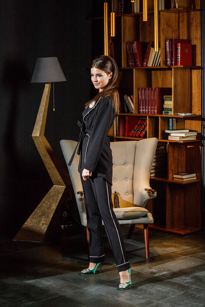 Portrait of beautiful brunette woman wearing pink t-shirt and black suit standing in home interior with bookshelf on background  - Φωτογραφία, εικόνα