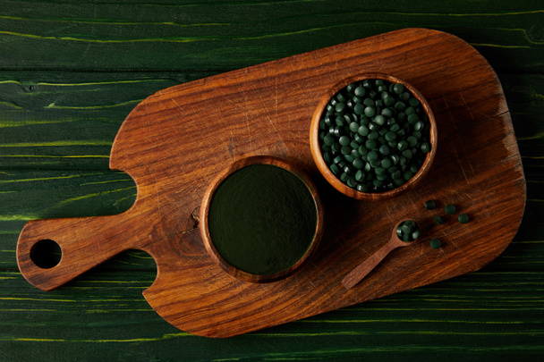 top view of cutting board with wooden spoon, bowls with spirulina powder and spirulina in pills on green wooden table  - Photo, Image