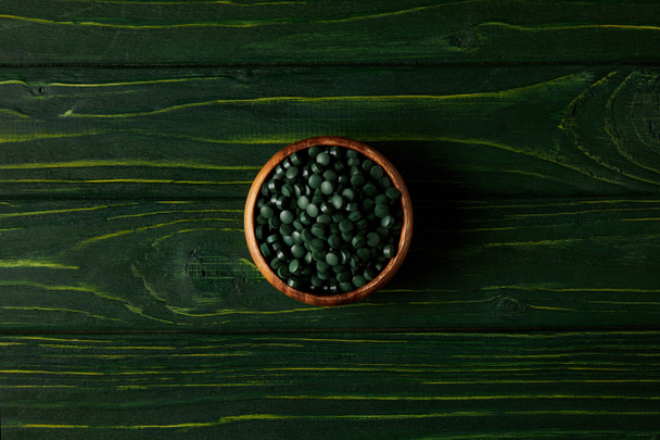 top view of wooden bowl with pile of spirulina pills on green wooden table - Fotó, kép