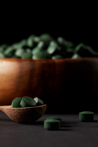 selective focus of wooden spoon and bowl with pile of spirulina pills on black background  - Photo, Image