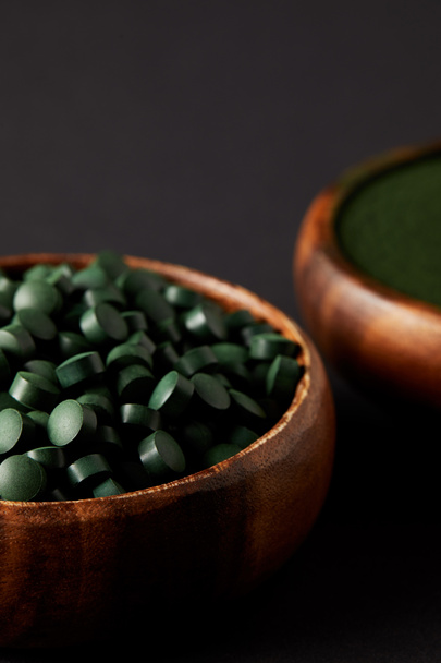 selective focus of wooden bowls with spirulina powder and spirulina in pills on grey background - Foto, afbeelding