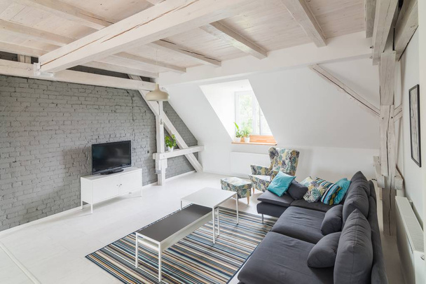 Φωτεινό σύγχρονο σαλόνι με τηλεόραση σύνολο και ύφασμα καναπέ. Γκρι τοίχο από τούβλα και ξύλινα δοκάρια στην οροφή. Μοντέρνα, φωτεινή σοφίτα διαμέρισμα. - Φωτογραφία, εικόνα