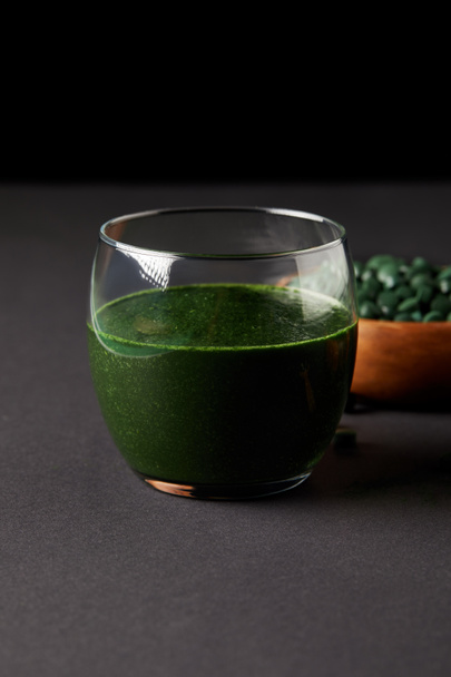 glass of fresh smoothie from spirulina and wooden bowl with spirulina pills on black background  - Fotografie, Obrázek