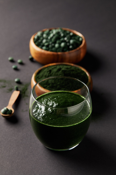 selective focus of arranged wooden spoon, glass of spirulina smoothie, bowls with spirulina powder and spirulina pills on grey table  - Photo, Image