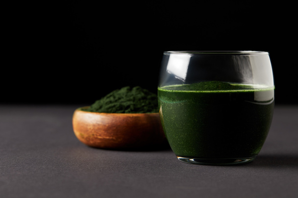 close up view of fresh spirulina drink in glass and spirulina powder in wooden bowl on black background  - 写真・画像