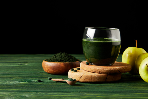 selective focus of wooden slices with fresh spirulina drink in glass, apples, wooden spoon and spirulina powder in wooden bowl on black background on black background  - 写真・画像