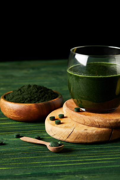 wooden slices with fresh spirulina drink in glass, wooden spoon and spirulina powder in wooden bowl on black background on green wooden table  - Foto, imagen