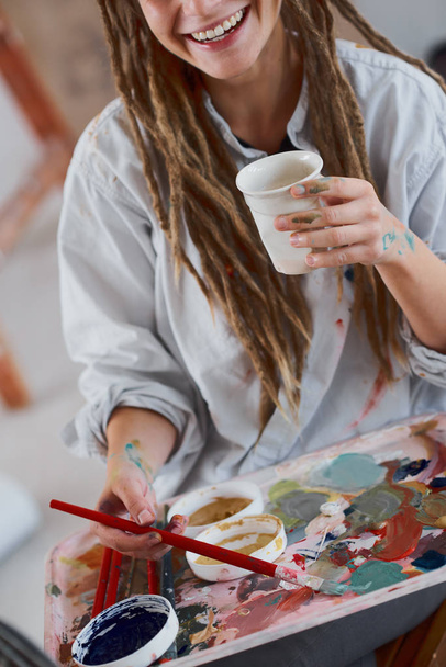 コーヒーを飲みながらスタジオで座っているとイーゼルの絵画芸術の女の子. - 写真・画像
