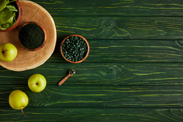 top view of cutting board, wooden bowls, spoon, apples, leaves, spirulina powder and spirulina pills on green wooden table  - Photo, Image