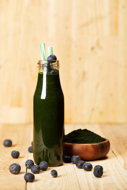 bowl with spiruluna powder, bottle of spirulina smoothie with blueberries and drinking straw on wooden table - Fotoğraf, Görsel