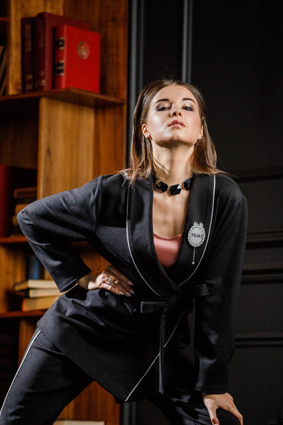 Portrait of beautiful brunette woman wearing pink t-shirt and black suit standing in home interior with bookshelf on background  - Zdjęcie, obraz