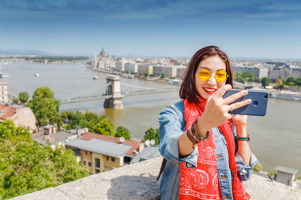 Happy young asian woman tourist making selfie photo by using her smartphone standing in front of the old bridge in Budapest city with view of the Parliament Building - 写真・画像