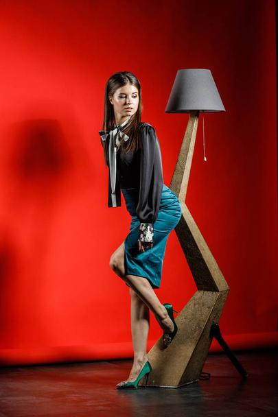 Studio portrait of young model in casual clothes on red background. Young woman in black shirt and blue skirt leaning on stylish wooden lamp at studio interior  - Фото, зображення