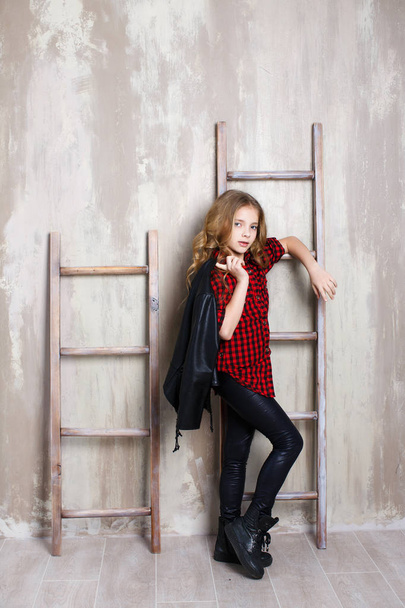 Портрет девочки-подростка с длинными волнистыми волосами светлого цвета, серыми глазами, одетой в красно-черную клетчатую рубашку и черные джинсы, на плече держит черную кожаную куртку. Студийное фото девушки, стоящей рядом с лестницей на сером фоне
 - Фото, изображение