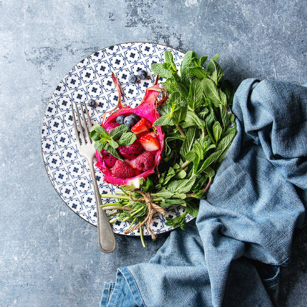Vegan fruit salad with berries and mint served in pink dragon fruit on ceramic plate with denim cloth over blue texture background. Top view, space. Square image - Fotografie, Obrázek