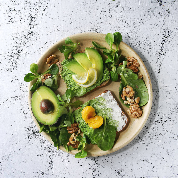Vegetarian sandwiches with avocado, ricotta, egg yolk, spinach, walnuts on whole grain toast bread on ceramic plate with ingredients above over white marble background. Top view, space - Foto, Imagen