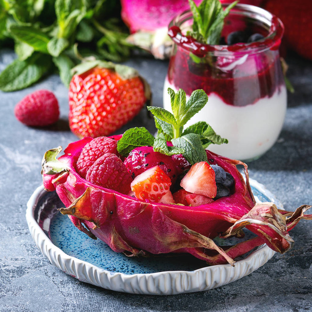 Vegan fruit salad with berries and mint served in pink dragon fruit with ingredients above on ceramic plate over blue texture background. Close up. Healthy eating - Foto, immagini