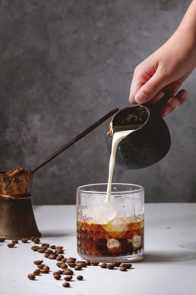 Iced coffee cocktail or frappe with ice cubes and cream, pouring from jug, served in glass with vintage jezva and coffee beans around on white marble table with grey concrete wall at background. - Fotografie, Obrázek