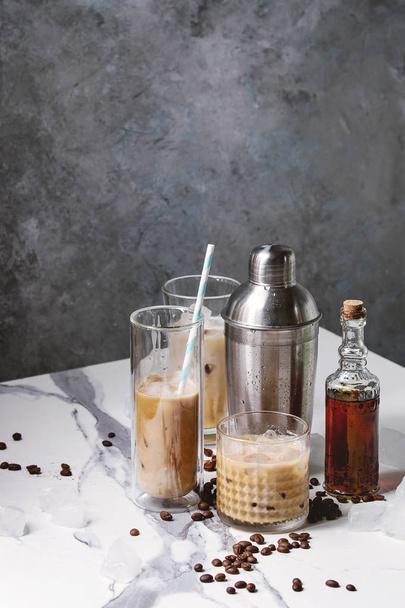 Iced coffee cocktail or frappe with ice cubes and cream in different glasses with silver shaker, bottle of rum, coffee beans around on white marble table with grey concrete wall at background. - Photo, Image