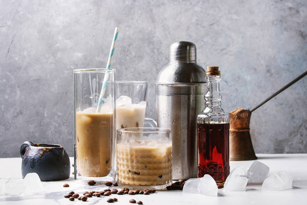 Iced coffee cocktail or frappe with ice cubes and cream in different glasses with jezva, silver shaker, bottle of rum, coffee beans around on white marble table with grey concrete wall at background. - Foto, Imagem