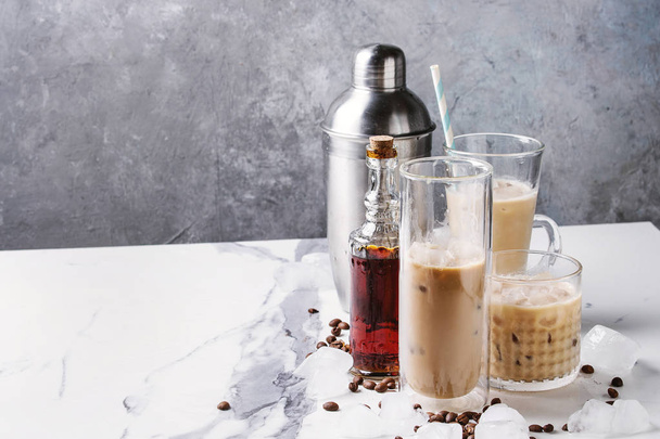 Iced coffee cocktail or frappe with ice cubes and cream in different glasses with silver shaker, bottle of rum, coffee beans around on white marble table with grey concrete wall at background. - Photo, image
