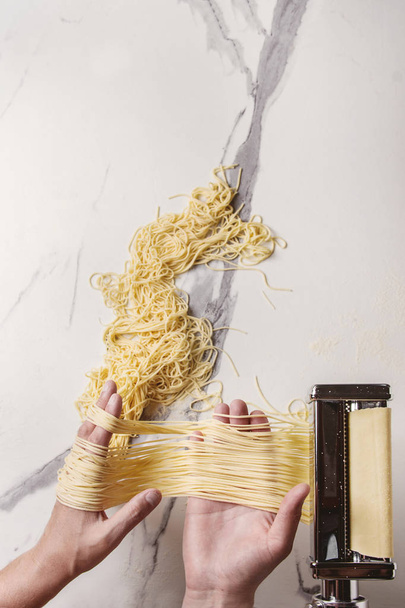 Rolled dough for homemade italian uncooked pasta spaghetti from pasta machine maker in hands with semolina flour over white marble texture background. Flat lay, space. - Photo, image