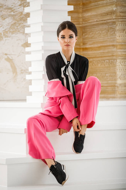 Studio portrait of beautiful brunette woman wearing pink suit and black t-shirt, sitting on stairs at luxury home interior  - Foto, imagen