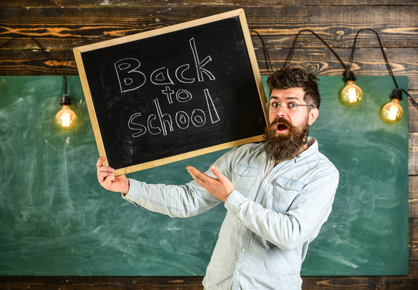 High school concept. Teacher in eyeglasses holds blackboard with title back to school. Man with beard and mustache on surprised face welcomes students, chalkboard on background - Фото, изображение