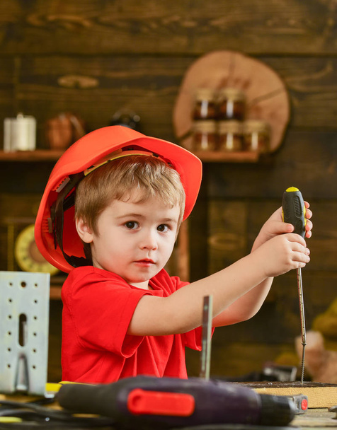 Boy in orange helmet and red T-shirt busy working. Concentrated kid binding screw into wood. Little carpenter playing with screwdriver - Foto, immagini