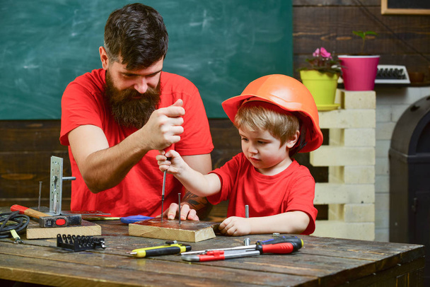 Teamwork in workshop concept. Father, parent with beard teaching little son to use tool screwdriver. Boy, child busy in protective helmet learning to use screwdriver with dad - Photo, image