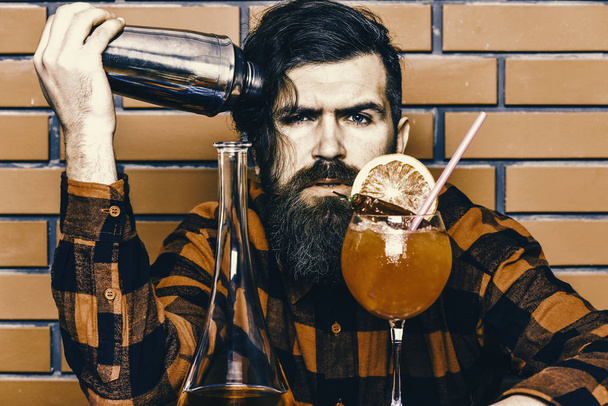 Man in checkered shirt holds shaker, brick wall background. Barman and cocktails concept. Barman with long beard and mustache and stylish hair with strict face near alcoholic cocktail, close up - Foto, Bild