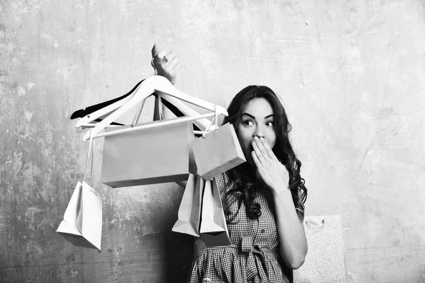 pretty sexy girl or cute fashionable woman with long brunette curly hair and smiling happy surprised face holds hangers, colorful shopping bags or packages on beige background - 写真・画像