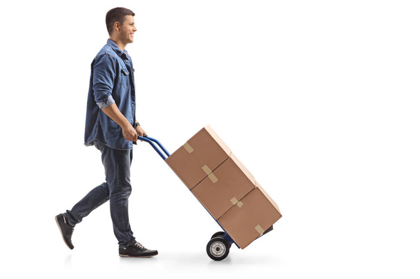Full length profile shot of a young man pushing a hand truck with boxes isolated on white background - Zdjęcie, obraz