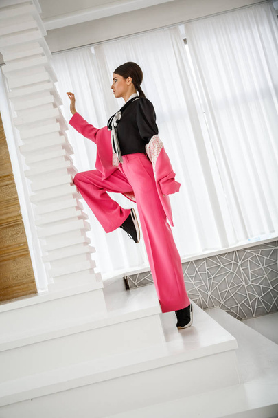 Studio portrait of beautiful brunette woman wearing pink suit and black t-shirt, standing on stairs at luxury home interior  - Zdjęcie, obraz