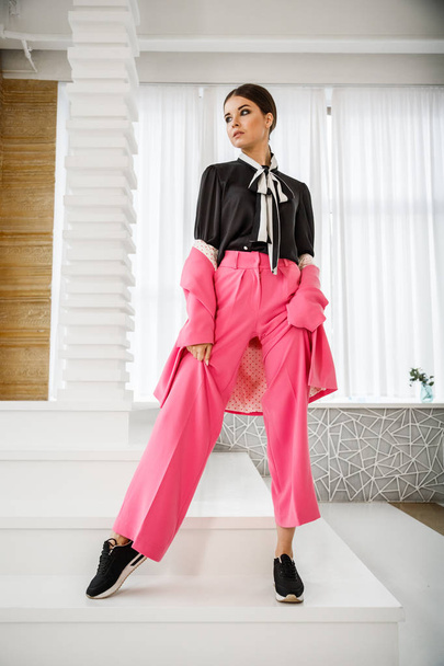 Studio portrait of beautiful brunette woman wearing pink suit and black t-shirt, standing on stairs at luxury home interior  - 写真・画像