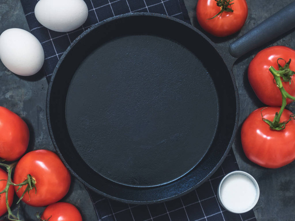 Black cast-iron frying pan on black cloth, white chicken eggs, red tomatoes and salt. Black background with blank space for text. Top view. Copy space. - Foto, Imagen