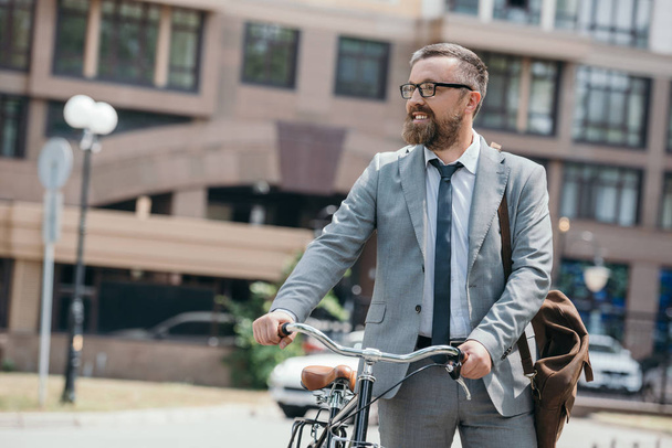 handsome businessman walking with bicycle on street in city and looking away - Photo, Image