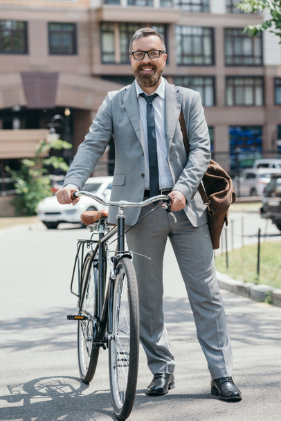 happy handsome businessman standing with bicycle and looking at camera on street in city - Foto, Imagem