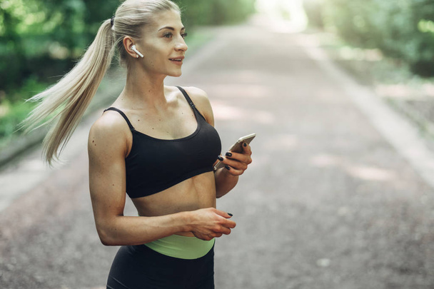 Woman Runner en el Summer Morning Park Escuchando música en Smartphone usando auriculares Bluetooth. Chica Fitness Femenina trotando en el camino fuera
. - Foto, imagen