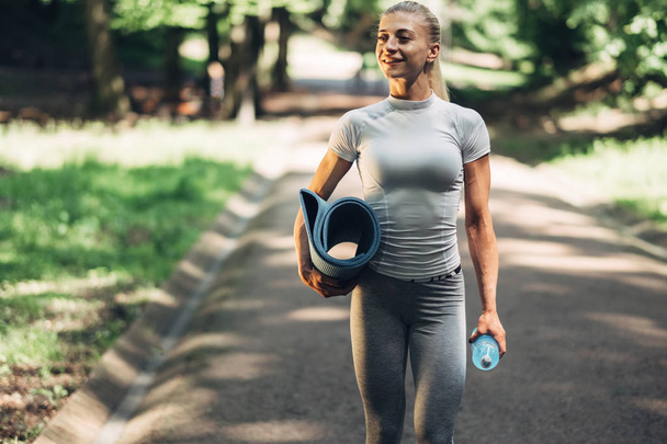 Fitness vrouw voorbereidingen voor Yoga in de vroege ochtend Park. Het dragen van stijlvolle Sport Outfit. Blauwe fles Water en Yoga Mat. Gezonde levensstijl. - Foto, afbeelding