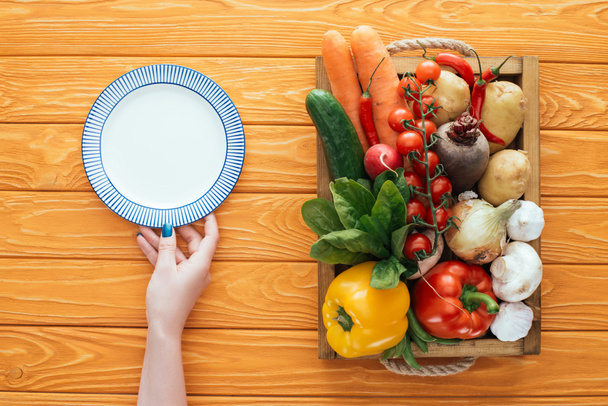 cropped shot of person holding empty round plate and fresh raw vegetables in box on wooden table top - Photo, Image