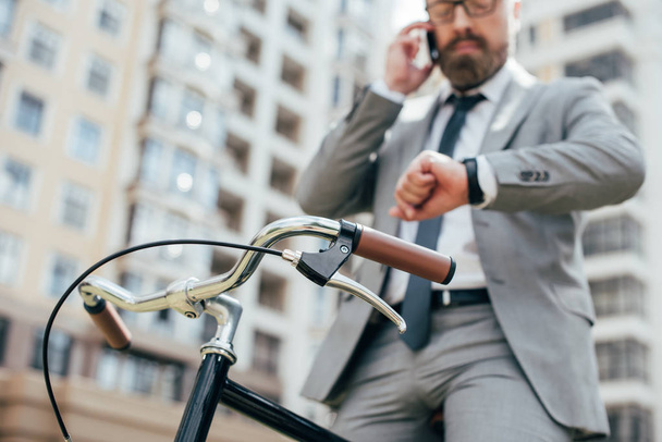 enfoque selectivo de hombre de negocios hablando en el teléfono inteligente y mirando reloj de pulsera mientras está sentado en bicicleta
  - Foto, imagen