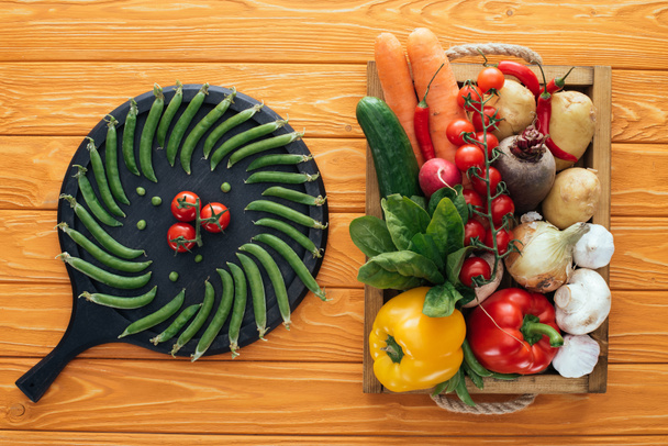 frische grüne Erbsen und Tomaten auf rundem Brett und rohes gesundes Gemüse in Schachtel auf hölzerner Tischplatte    - Foto, Bild