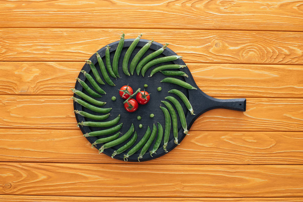 bovenaanzicht van verse groene erwten en tomaten op ronde bord op houten tafelblad   - Foto, afbeelding
