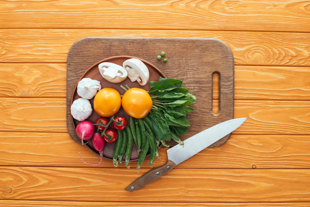 top view of fresh raw vegetables in plate on wooden cutting board and knife on wooden table top - Photo, Image