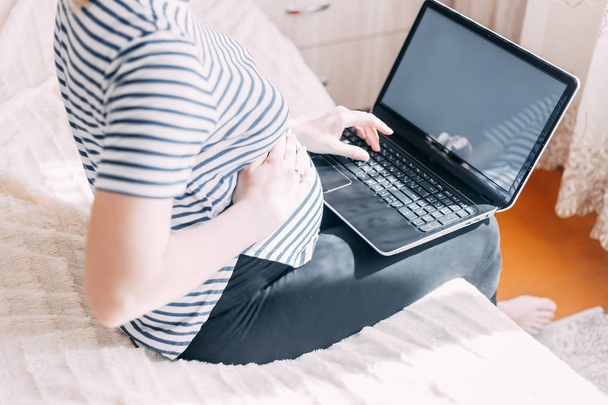 妊娠中の女性のラップトップに取り組んでいます。彼女の仕事場を自宅で座っている間ノート パソコンに何かを入力する妊娠中の実業家の画像をトリミング - 写真・画像