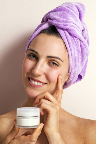 photo of young woman in purple towel on head holding face cream on pink background - Фото, изображение