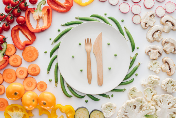 top view of wooden fork and knife on round white plate and fresh organic vegetables isolated on white - Zdjęcie, obraz