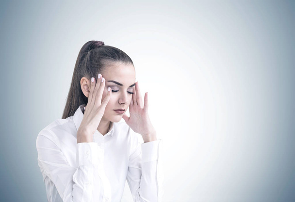 Stressed young businesswoman with long hair in a ponytail wearing a white shirt. She is having a headache and standing with closed eyes. A mock up gray wall background - Foto, Bild
