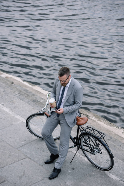 businessman with coffee to go using smartphone and standing with bike on quay - Photo, Image